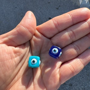 May include: Two small, square-shaped, blue glass evil eye beads. One bead is a light blue color and the other is a darker blue color. Both beads have a white evil eye design in the center.