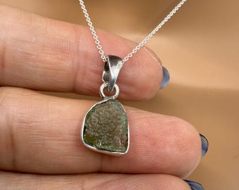 May include: A silver necklace featuring a pendant with a rough-cut, olive-green gemstone. The pendant is set in a silver bezel and hangs from a delicate silver chain. The necklace is displayed against a neutral background.