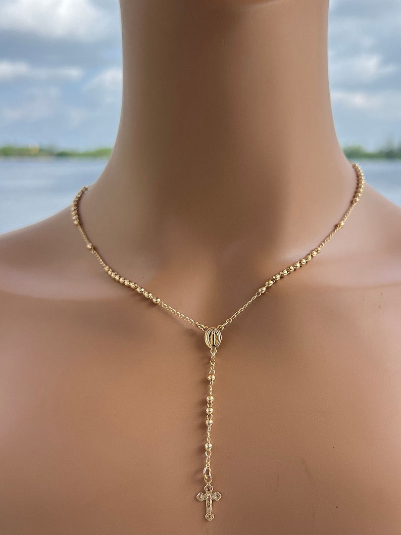 Puede incluir: Un collar de rosario dorado con un peque&ntilde;o dije de cruz y un dije redondo.
