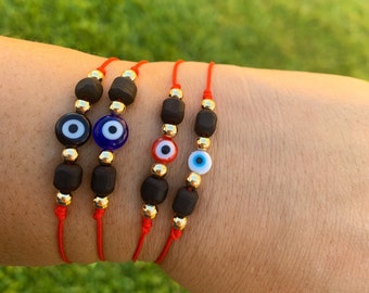 Puede incluir: Cuatro pulseras de hilo rojo con cuentas de madera, detalles dorados y amuletos de ojo turco. Los amuletos de ojo turco son de color negro, azul, rojo y blanco. Las pulseras se muestran sobre un fondo verde.