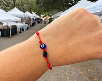 May include: Red string bracelet featuring a blue and white evil eye bead and a black geometric bead. The bracelet is knotted and designed to be worn on the wrist. The background shows an outdoor market with white tents and green trees.