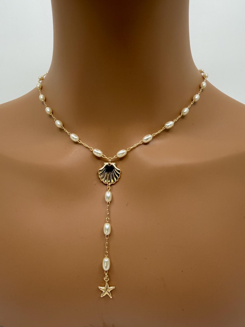May include: Gold-toned necklace featuring a seashell pendant with dangling pearls and a starfish charm. The necklace is adorned with small, oval-shaped pearls along the chain, creating a delicate and elegant design.