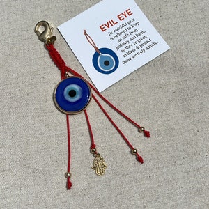 May include: A red string charm with a blue evil eye charm and a gold hamsa hand charm. The evil eye charm is round and has a white pupil. The hamsa hand charm is a cutout design. The string is tied with knots and has gold beads. The charm is believed to protect against evil and bring good luck.