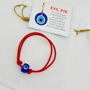 May include: Red string bracelet with a blue evil eye charm. The bracelet comes in a small white drawstring bag with a card that reads "Evil Eye. Its watchful gaze is believed to keep us safe from jealousy and harm, so they're given to bless & protect those we truly admire."