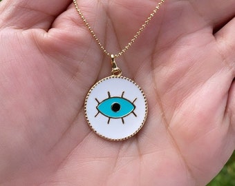 May include: Gold-toned necklace featuring a circular pendant with a white background and a turquoise eye design. The eye has a black pupil and is surrounded by black lines. The pendant is held by a delicate gold chain.