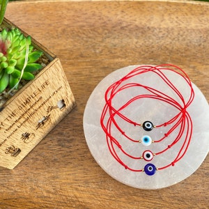 May include: Five red string bracelets with different colored evil eye charms. The charms are blue, white, black, and red. The bracelets are on a white circular surface.