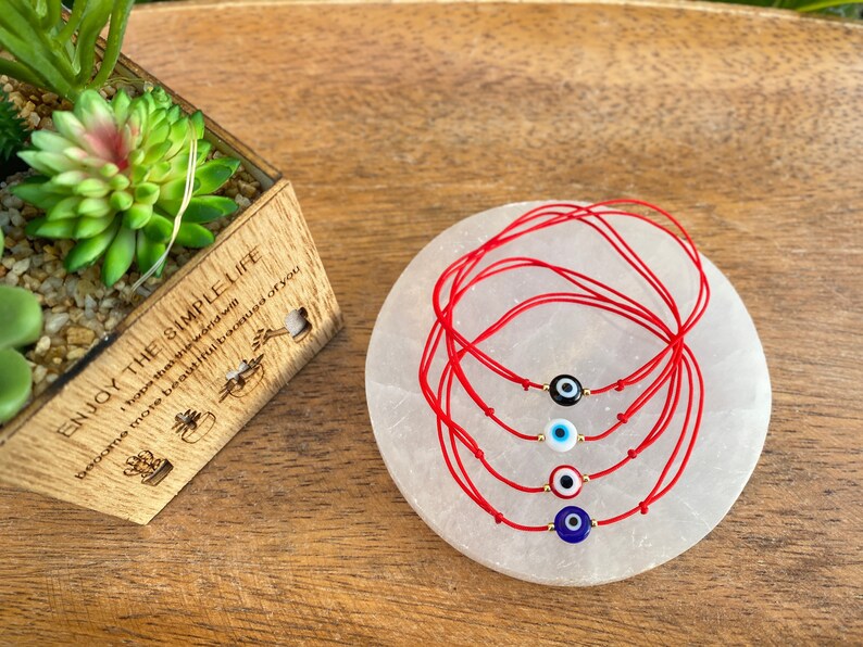 May include: Five red string bracelets with different colored evil eye charms. The charms are blue, white, black, and red. The bracelets are on a white circular surface.