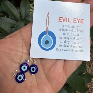 May include: A hand holding three small blue evil eye charms with silver-colored findings. A card with the text "EVIL EYE" and a description of the charm's purpose is also visible. The charms are a deep blue with a white and black eye design.