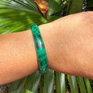 May include: A smooth, cylindrical bangle bracelet featuring a vibrant green malachite pattern with characteristic dark green swirling bands and concentric circular markings throughout the polished surface of the circular wrist accessory.