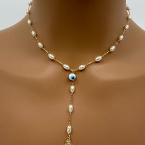 May include: Gold-toned necklace featuring a delicate chain adorned with small, oval pearls. The necklace includes a blue and white evil eye charm and a gold Hamsa hand pendant, creating a unique and stylish accessory.