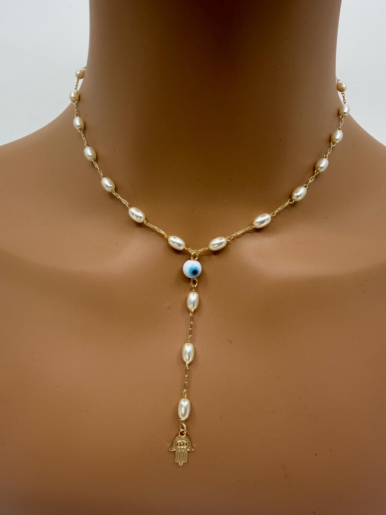 May include: Gold-toned necklace featuring a delicate chain adorned with small, oval pearls. The necklace includes a blue and white evil eye charm and a gold Hamsa hand pendant, creating a unique and stylish accessory.