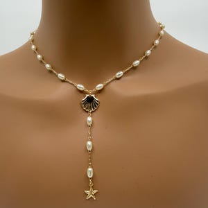 May include: Gold-toned necklace featuring a seashell pendant with a dangling strand of pearls and a starfish charm. The necklace has a delicate chain with small pearls. The seashell pendant has black accents.