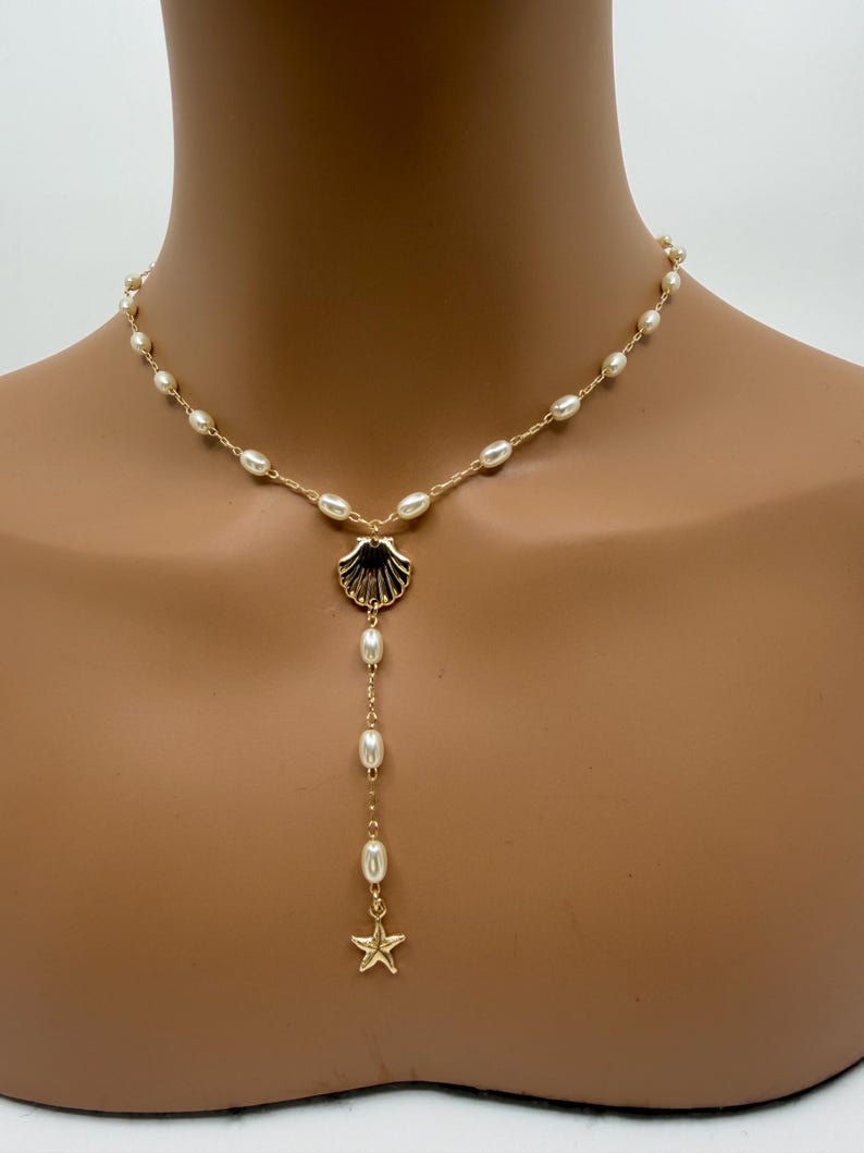 May include: Gold-toned necklace featuring a seashell pendant with a dangling strand of pearls and a starfish charm. The necklace has a delicate chain with small pearls. The seashell pendant has black accents.