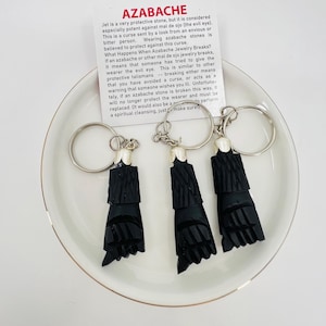 May include: Three black azabache keyrings with silver key rings and white details are displayed on a white plate. A card with text about azabache is also present. The keyrings are carved in a stylized shape.