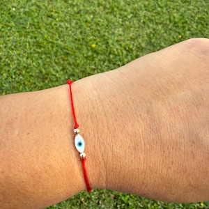 May include: Red string bracelet featuring a white and blue evil eye charm with gold-colored beads. The bracelet is a simple design, with the charm as the focal point. The background is green grass.