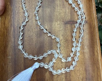 Op de afbeelding: Een ketting van heldere kwartskristallen kralen met een witte kwast. De kralen zijn rond en geregen aan een wit koord. De kwast is gemaakt van witte draden en is bevestigd aan het uiteinde van de ketting.