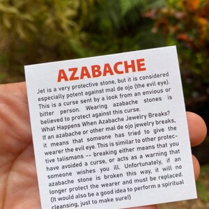 May include: A white card with black text explaining the properties of azabache, a protective stone. The text reads "Azabache Jet is a very protective stone, but it is considered especially potent against mal de ojo (the evil eye). This is a curse sent by a look from an envious or bitter person. Wearing azabache stones is believed to protect against this curse. What Happens When Azabache Jewelry Breaks? If an azabache or other mal de ojo jewelry breaks, it means that someone has tried to give the wearer the evil eye. This is similar to other protective talismans -- breaking either means that you have avoided a curse, or acts as a warning that someone wishes you ill. Unfortunately, if an azabache stone is broken this way, it will no longer protect the wearer and must be replaced. (It would also be a good idea to perform a spiritual cleansing, just to make sure!)"