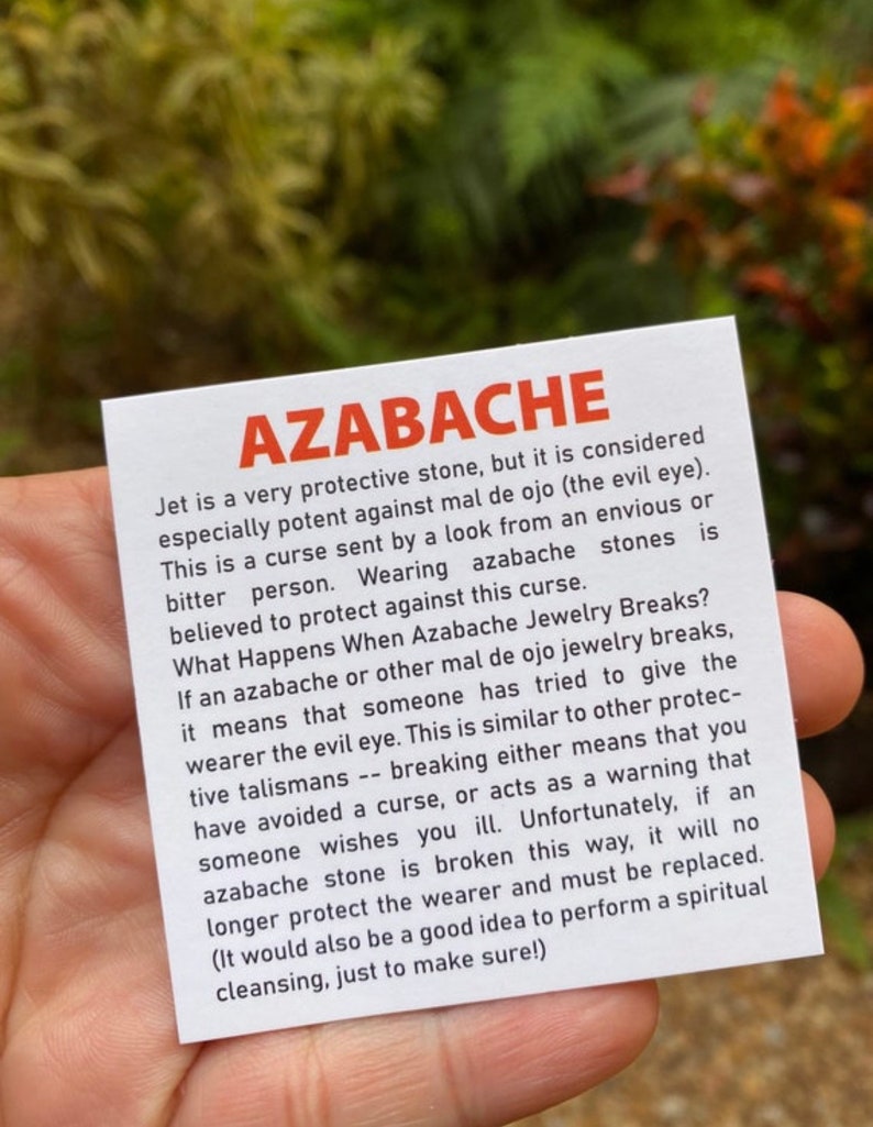 May include: A white card with black text explaining the properties of azabache, a protective stone. The text reads "Azabache Jet is a very protective stone, but it is considered especially potent against mal de ojo (the evil eye). This is a curse sent by a look from an envious or bitter person. Wearing azabache stones is believed to protect against this curse. What Happens When Azabache Jewelry Breaks? If an azabache or other mal de ojo jewelry breaks, it means that someone has tried to give the wearer the evil eye. This is similar to other protective talismans -- breaking either means that you have avoided a curse, or acts as a warning that someone wishes you ill. Unfortunately, if an azabache stone is broken this way, it will no longer protect the wearer and must be replaced. (It would also be a good idea to perform a spiritual cleansing, just to make sure!)"
