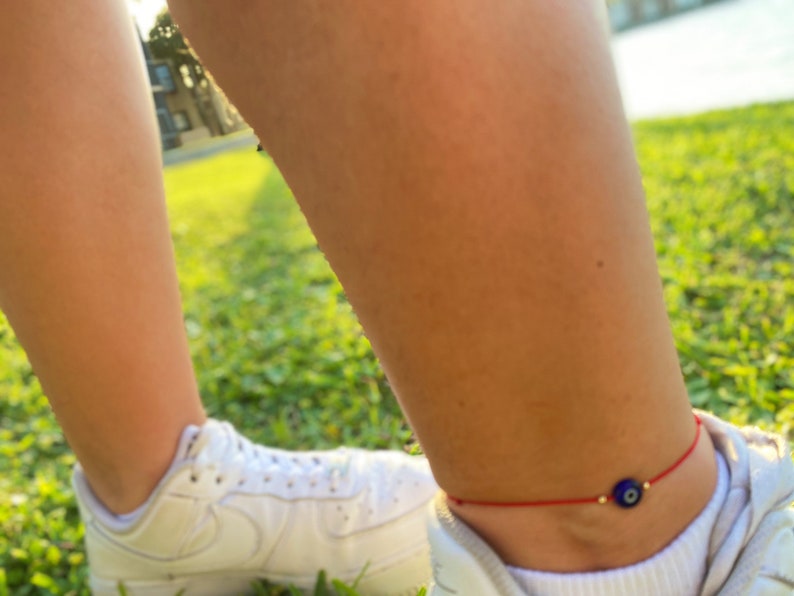May include: A red string bracelet with a blue evil eye charm. The bracelet is worn on a person's ankle.