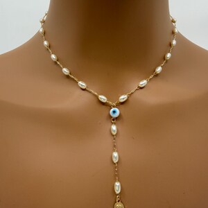 May include: A gold-toned necklace featuring a strand of small, white pearls. The necklace has a drop pendant with a blue and white evil eye bead, more pearls, and a gold-colored Hamsa hand charm.