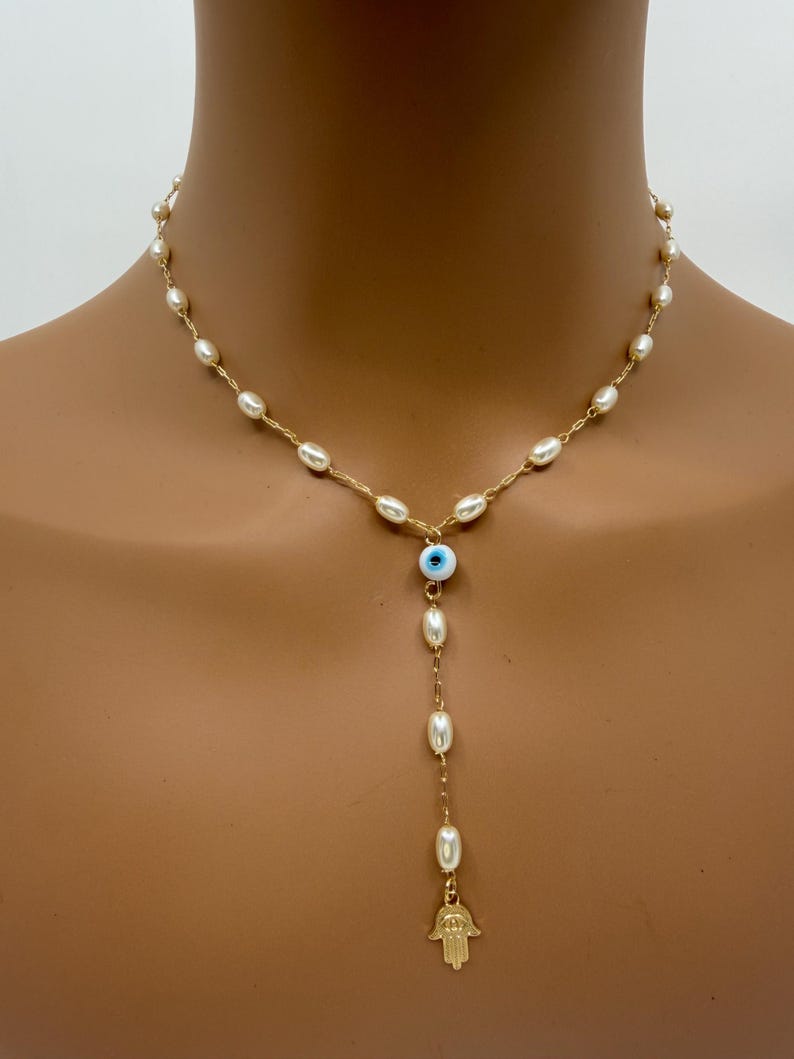 May include: A gold-toned necklace featuring a strand of small, white pearls. The necklace has a drop pendant with a blue and white evil eye bead, more pearls, and a gold-colored Hamsa hand charm.