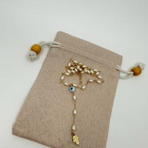 May include: A gold-colored necklace with white oval-shaped beads and a blue eye charm, resting on a beige drawstring pouch. The necklace has a small gold-colored Hamsa hand pendant at the end. The pouch has wooden beads on the drawstrings.