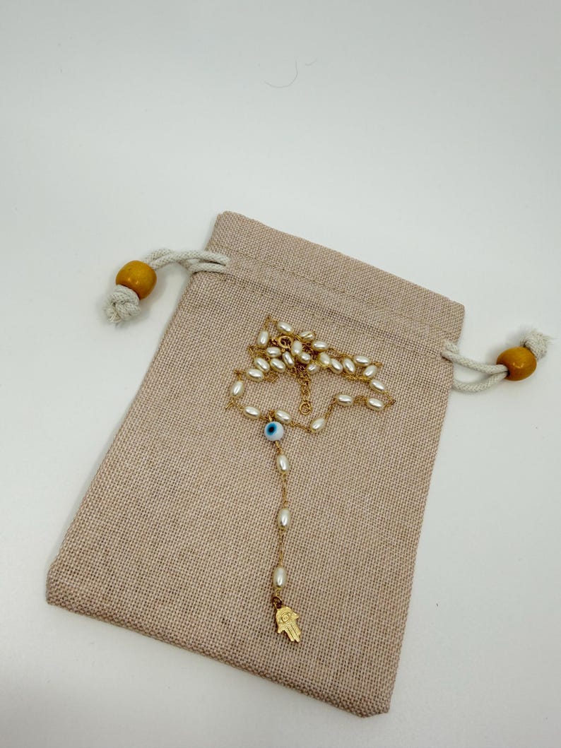 May include: A gold-colored necklace with white oval-shaped beads and a blue eye charm, resting on a beige drawstring pouch. The necklace has a small gold-colored Hamsa hand pendant at the end. The pouch has wooden beads on the drawstrings.