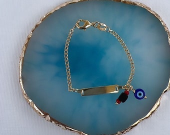 May include: A gold-colored bracelet with a rectangular nameplate and a chain. The bracelet features a charm with a blue eye, red beads, and black beads. The bracelet is displayed on a blue and gold-edged surface.