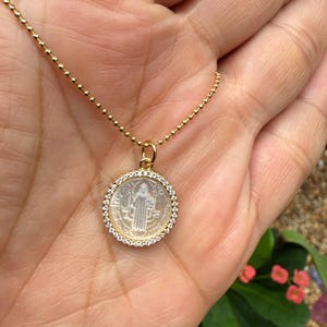 Puede incluir: Un collar dorado con un colgante redondo. El colgante presenta una figura religiosa rodeada de pequeñas piedras transparentes. La cadena está hecha de pequeñas cuentas redondas.