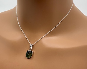 May include: A silver necklace with a pendant featuring a rough-cut, dark green gemstone. The pendant is set in a silver bezel and hangs from a delicate silver chain. The necklace is displayed on a person's neck.