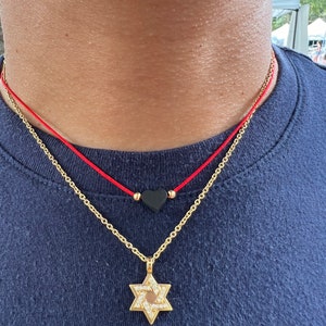 May include: A gold chain necklace with a diamond-encrusted Star of David pendant. A red string necklace with a black heart charm is layered on top.