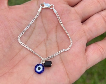 Puede incluir: Un brazalete de cadena de plata con un dije de ojo turco azul y una cuenta negra.
