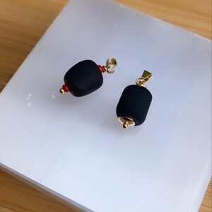 May include: Two black pendant charms with gold accents. The charms are shaped like a cylinder and have a small red bead on one end.
