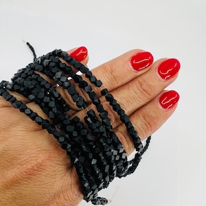 May include: A strand of black faceted beads, likely made of glass or gemstone, is shown on a hand. The beads are small and have a uniform shape.