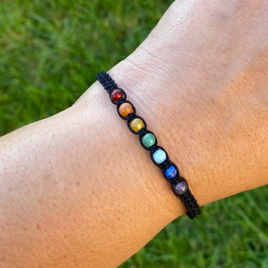 May include: A black string bracelet with seven colourful beads. The beads are red, orange, yellow, green, blue, indigo, and violet. The bracelet is a chakra bracelet.