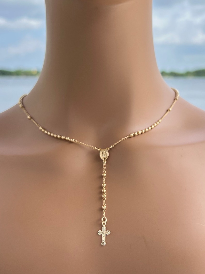 Puede incluir: Un collar de rosario dorado con un peque&ntilde;o colgante de cruz. El collar est&aacute; hecho de peque&ntilde;as cuentas redondas.