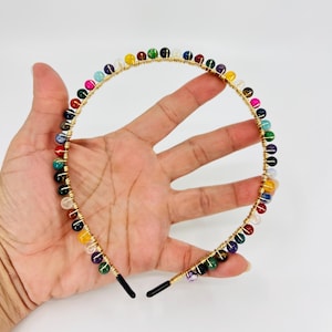 Puede incluir: Una diadema dorada con un alambre envuelto alrededor y decorada con cuentas de piedras preciosas de colores.