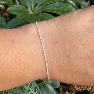 May include: A delicate gold bracelet with a line of small, sparkling stones. The bracelet is thin and elegant, perfect for everyday wear. The bracelet is shown on a wrist against a backdrop of green foliage.