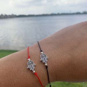 May include: Two bracelets with Hamsa hand charms. One bracelet has a red cord, and the other has a black cord. The Hamsa hand charms are silver-colored and detailed. The bracelets are worn on a person's wrist.