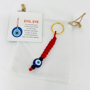 May include: Red string keychain with a blue evil eye charm. The keychain is in a white drawstring bag. The evil eye is believed to protect against jealousy and harm. "Its watchful gaze is believed to keep us safe from jealousy and harm, so they're given to bless & protect those we truly admire."