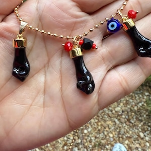 Pode incluir: Um colar de corrente dourada com três pingentes de punho preto, cada um com uma tampa dourada. O colar também inclui contas vermelhas e pretas e um amuleto de olho grego azul e branco. O colar é exibido em uma mão.