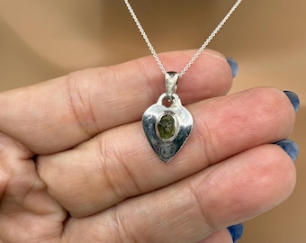 May include: A silver pendant necklace featuring a heart-shaped pendant with a green gemstone. The pendant hangs from a delicate silver chain. The necklace is displayed against a neutral background.