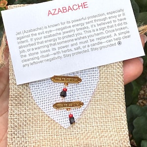 May include: A small burlap pouch with a white fabric insert, holding two pairs of earrings. Each earring has a black stone and a red bead. A card with the word "AZABACHE" and text about protection is attached.