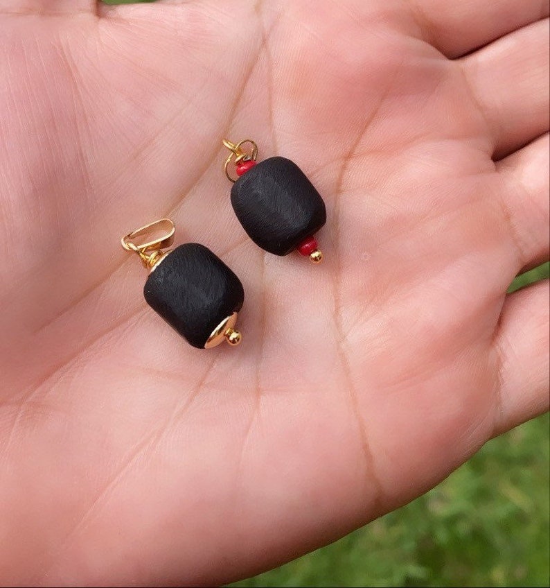 May include: Two black wood bead pendants with gold accents and red beads. The pendants are on a hand.