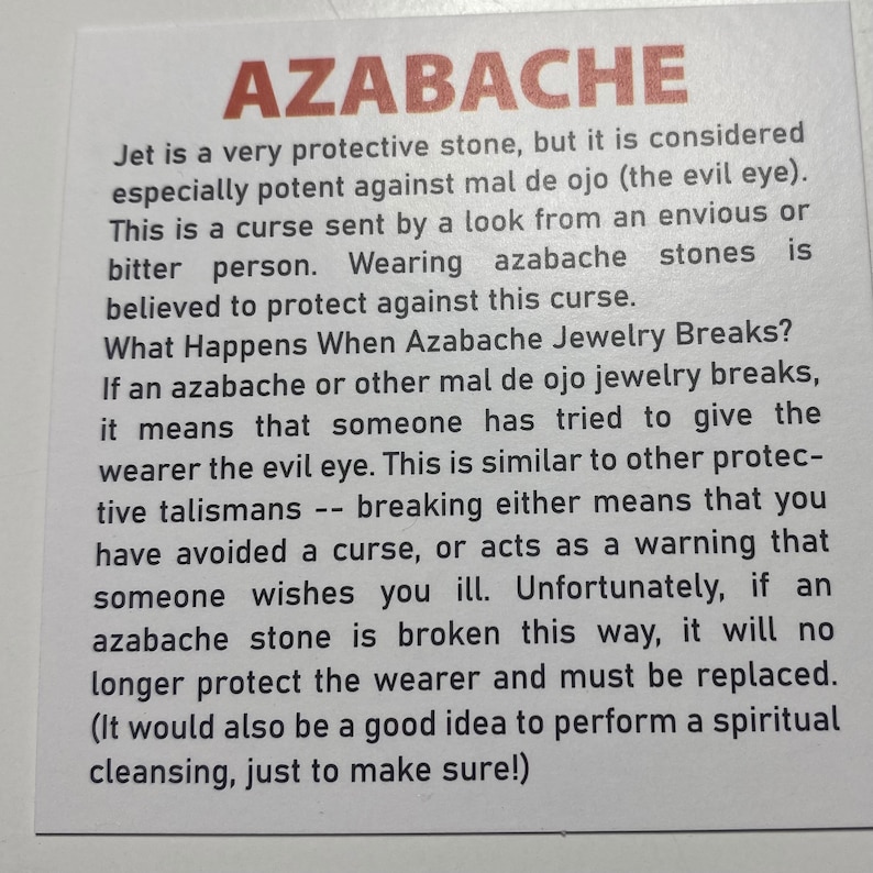 May include: A close-up of a white card with black text explaining the meaning of an azabache stone, a protective stone believed to ward off the evil eye. The text explains that if the stone breaks, it means someone has tried to give the wearer the evil eye. The text also states that the stone should be replaced if it breaks.