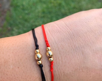 May include: Two bracelets with gold beads on red and black string. The bracelets are tied around a person's wrist.