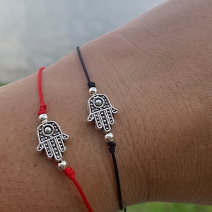 Puede incluir: Dos pulseras con dijes de mano de Hamsa plateados. Una pulsera tiene un cordón rojo y la otra un cordón negro.
