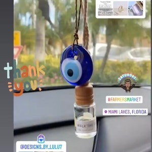 May include: A blue evil eye charm hanging from a rearview mirror with a small glass bottle of car diffuser that says "Walk the Walk Car Diffuser". The charm is attached to a string and the bottle is hanging from the string. The image also includes the text "@barbeite.co", "#farmersmarket", "Miami Lakes, Florida", and "@designs_by_lulu7".