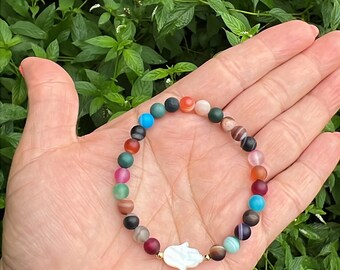 May include: A colourful beaded bracelet with a white hamsa charm. The bracelet is made of agate beads in various colours, including red, orange, yellow, green, blue, and purple.