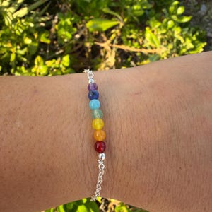 May include: A silver bracelet with seven coloured gemstone beads, representing the chakras. The bracelet is worn on a wrist, with green foliage in the background. The gemstones are red, orange, yellow, green, blue, indigo, and violet.